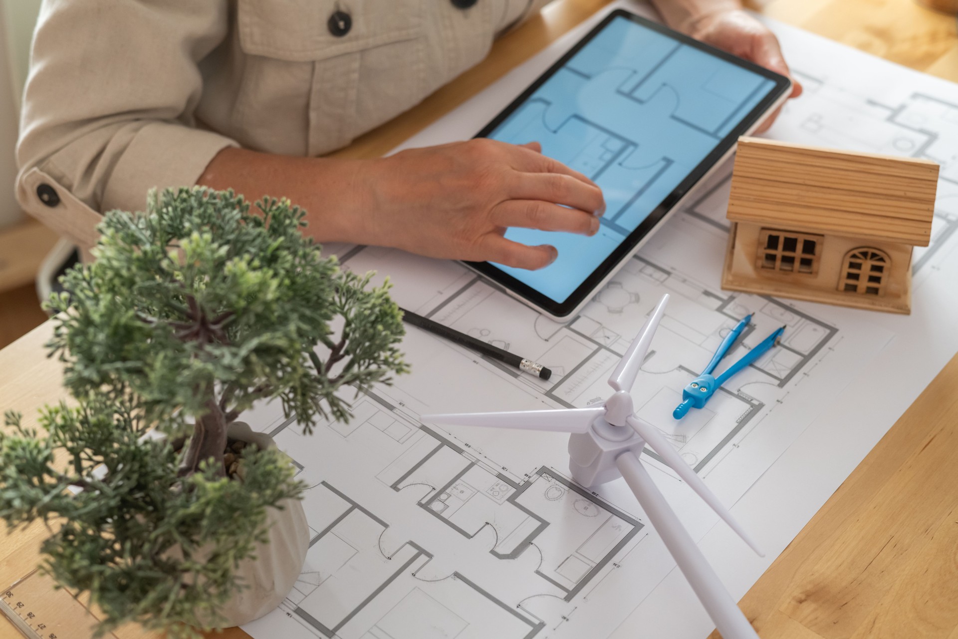 Architect is designing a sustainable and ecological house project, using a tablet and evaluating the blueprints with a miniature wind turbine and a small wooden house model on the desk